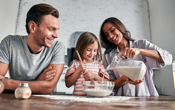 Cute little girl i jej pięknych rodziców są ugniatać mąki do pieczenia i uśmiech podczas gotowania w kuchni. - Zdjęcie, obraz