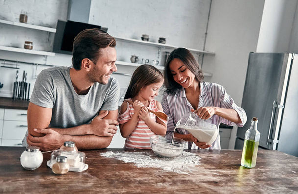 милая маленькая девочка и ее красивые родители месить муку для выпечки и улыбки во время приготовления пищи на кухне. - Фото, изображение