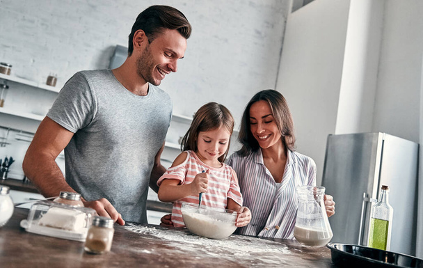 Cute little girl and her beautiful parents are knead flour for baking and smile while cooking in the kitchen. - Photo, Image