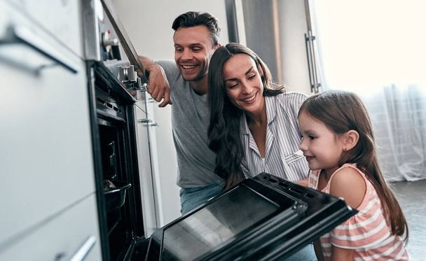 Happy young family with their little cute daughter are peep into the oven while cooking cakes on kitchen. - Foto, Imagem
