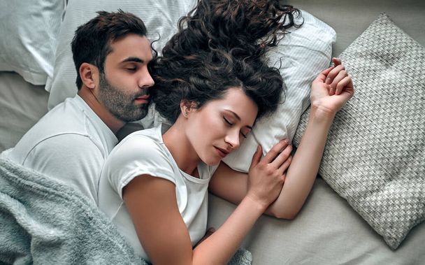 Top view of beautiful young couple hugging while sleeping together in bed at home - 写真・画像