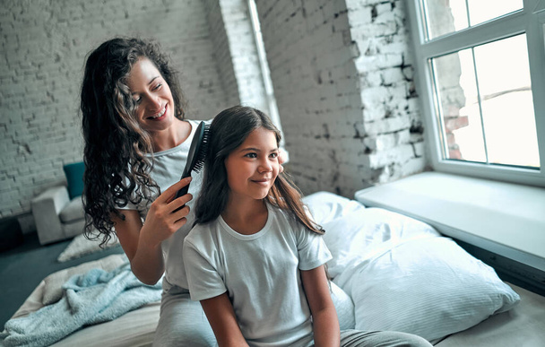 Hermosa madre joven está peinando su lindo pelo de hija pequeña. Ambos en pijama sentados en la cama y sonriendo - Foto, Imagen