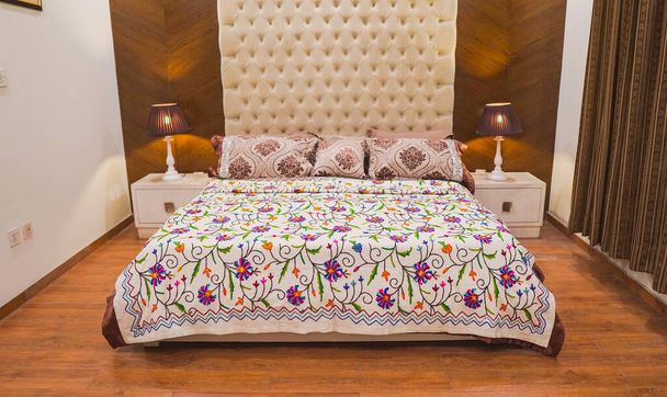 Eigentijdse Bed met paarse en groene bloem laken en enorme witte hoofdeinde en bruine achtergrond - Foto, afbeelding