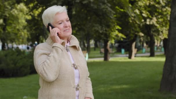 Feliz sereno senior 60s hermosa mujer caucásica abuela hablando en el teléfono celular moderno, maduro tranquilo 70s dama relajante al aire libre, hablar tienen llamada de teléfono inteligente agradable, ancianos y concepto de tecnología - Imágenes, Vídeo