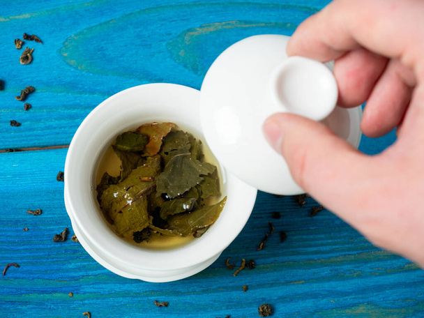 a bowl of brewed tea on a blue wooden background. A hand lifts the lid of the bowl. The opened leaves of large-leaved tea. Chinese tea. tea party - Fotó, kép