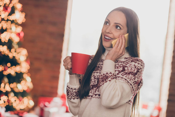 Photo of funny impressed young lady talk telephone drink tea near tree wear ugly sweater new year at home - Photo, image