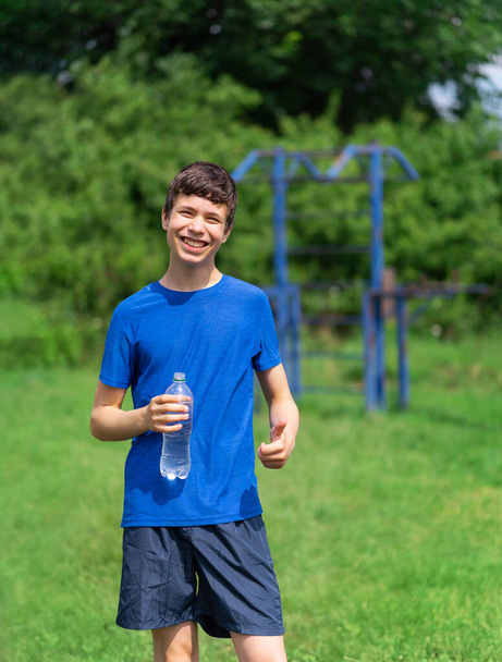 teenage boy exercising outdoors, sports ground in the yard, he opens a bottle of water and drinks, healthy lifestyle - Photo, Image