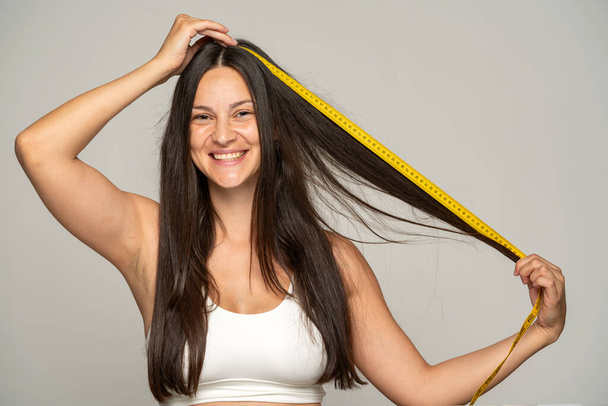 uma jovem mulher feliz mostra o comprimento de seu cabelo com uma fita métrica em um fundo cinza - Foto, Imagem