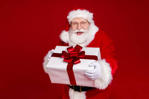 Photo of positive excited retired man wear santa claus costume spectacles smiling holding present box isolated red color background - Фото, изображение