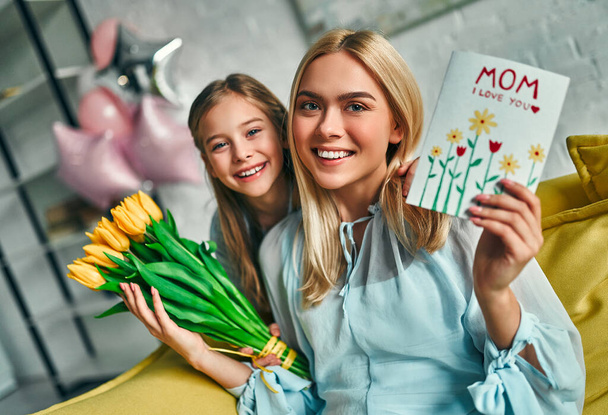 Happy mother's day! Child daughter congratulates mom and gives her flowers tulips and postcard. Mum and girl smiling and hugging. Family holiday and togetherness. - Foto, Imagen