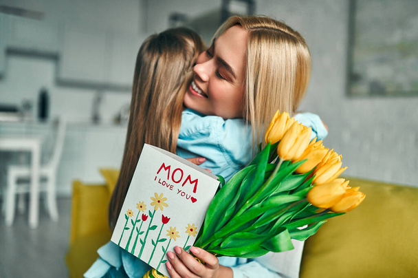 glücklicher Muttertag! Tochter gratuliert Mama und schenkt ihr Blumen, Tulpen und Postkarten. Mutter und Mädchen lächeln und umarmen sich. Familienurlaub und Zweisamkeit. - Foto, Bild