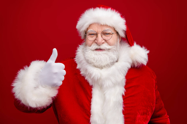 Photo of excited funny retired man wear santa claus costume spectacles smiling showing thumb up isolated red color background - Photo, image