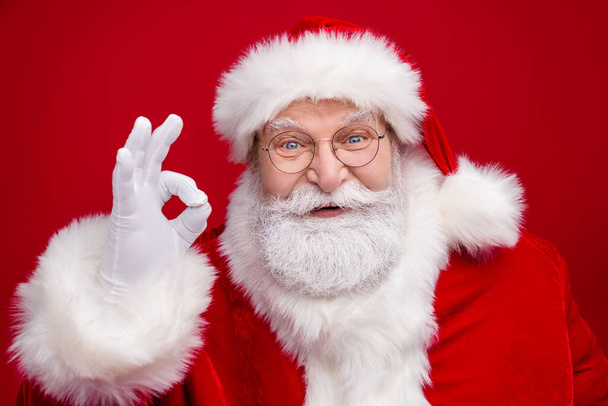 Photo of funny excited retired man wear santa claus costume spectacles smiling showing okey sign isolated red color background - 写真・画像