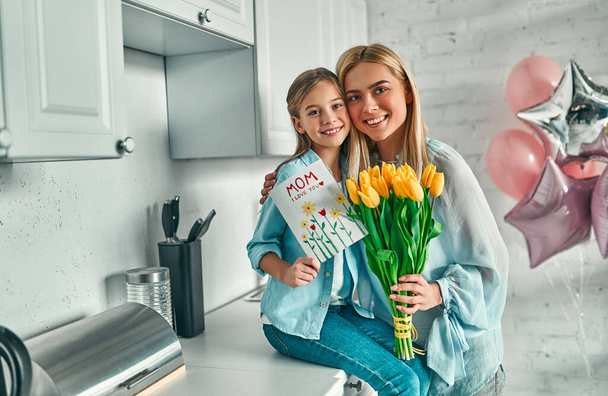 Gelukkige moederdag! Kind dochter feliciteert moeder en geeft haar bloemen tulpen en briefkaart. Mama en meisje glimlachend en knuffelen. Vakantie met het gezin en saamhorigheid. - Foto, afbeelding