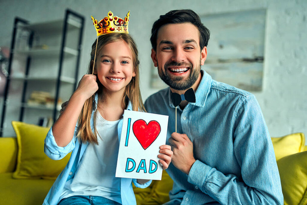 Funny family! Father and his child daughter with a paper accessories. Beauty funny girl holding paper crown on stick. She congratulate dady and gives him a postcard on Happy Father's Day! - Фото, зображення