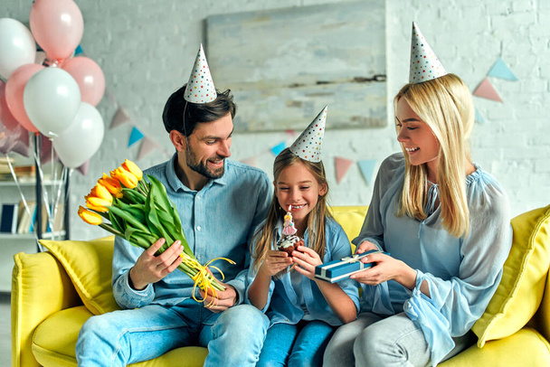 誕生日おめでとう！幸せな両親と娘。小さな女の子の誕生日のための家族とのパーティー、部屋は風船で飾られています。父と母は娘を祝福し、彼女に贈り物と花チューリップを与えます. - 写真・画像