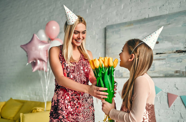 Happy birthday! Mother congratulates daughter and gives her flowers , pretty mom and lovely cute daughter wears similar festive dress and birthday hats. Family lovely moments. - Photo, Image