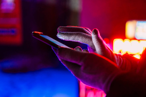 Großaufnahme von Frauenhänden in Winterhandschuhen, die ein Mobiltelefon vor dem Hintergrund von Neonlichtern auf der Straße halten, kopieren den Raum. Bezahlen im Online-Banking, Arbeiten in Anwendungen - Foto, Bild