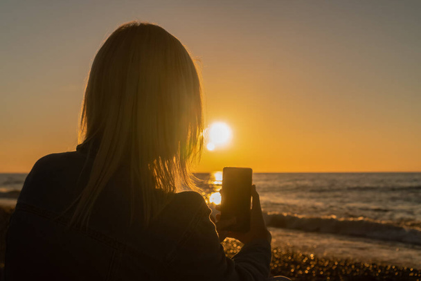 Young woman is sitting on a pebble beach by the sea and taking pictures on her mobile phone of a beautiful sunset, a photo against the sun. Concept of happiness, travel, lifestyle - Photo, image