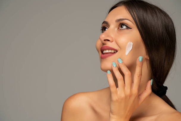 portrait of a young beautiful happy woman applies a skin product on her face on a gray background - Foto, Bild