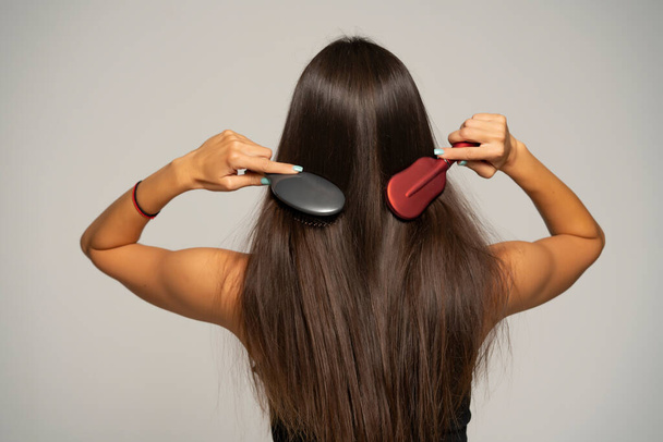 Вид сзади на женщину, расчесывающую волосы двумя расческами на сером фоне - Фото, изображение