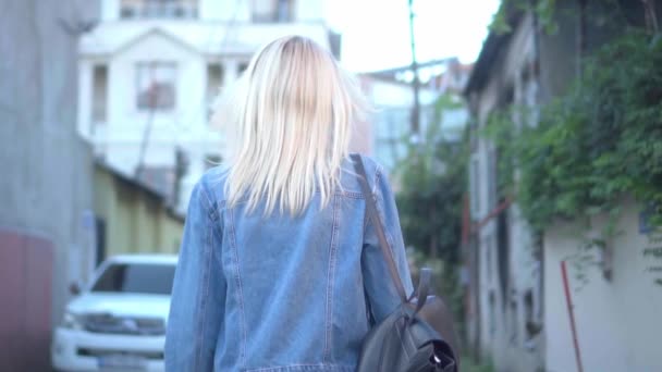 Achteraanzicht van jonge stijlvolle blonde in denim jas wandelen door het centrum - Video