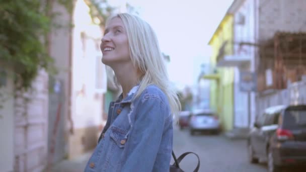 Вид сзади на молодую стильную блондинку в джинсовой куртке, идущую по центру города - Кадры, видео