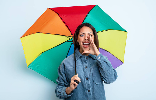 pretty hispanic woman feeling happy,giving a big shout out with hands next to mouth. umbrella concept - Fotografie, Obrázek