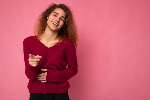 Young european happy positive charming cute brunette curly woman with sincere emotions wearing trendy pink sweater isolated over pink background with free space and laughing - Photo, Image
