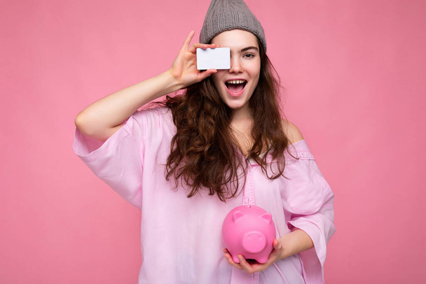 Viehättävä iloinen iloinen nuori brunette nainen yllään paita eristetty vaaleanpunainen tausta vapaata tilaa ja tilalla vaaleanpunainen sika rahaa laatikko ja luottokortti mockup - Valokuva, kuva