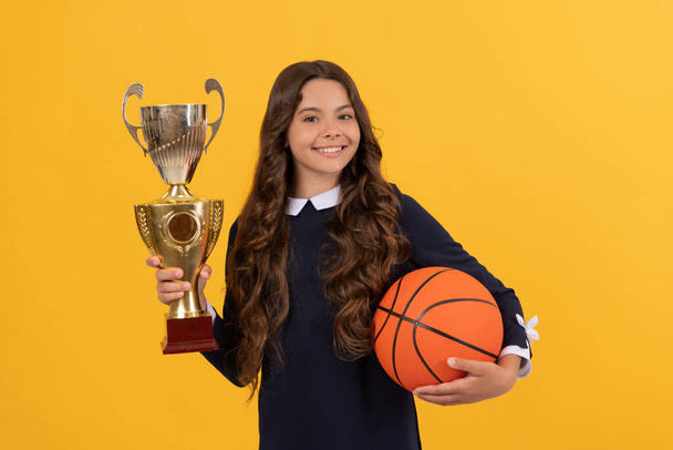 sonriente niño mantenga pelota de baloncesto y copa campeón sobre fondo amarillo, logro - Foto, imagen