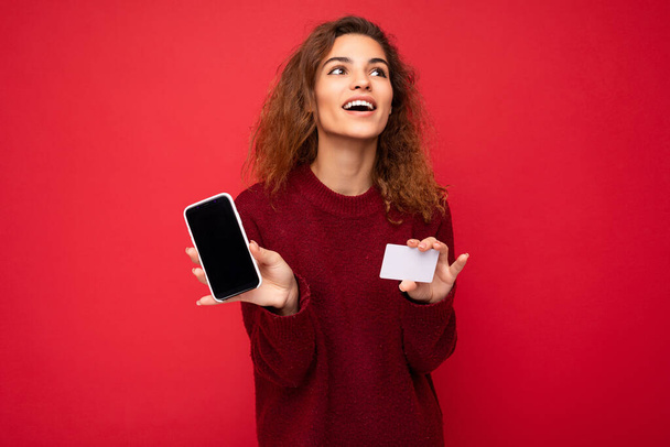 Fiatal gyönyörű göndör barna nő őszinte érzelmek elszigetelt háttér fal felett üres hely visel alkalmi sötét piros pulóver gazdaság hitelkártya mobiltelefon üres képernyőn - Fotó, kép