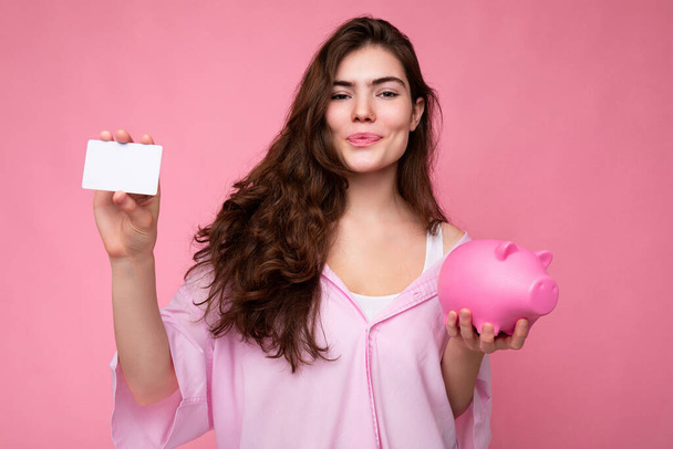 Attrayant jeune femme brune heureuse portant chemise isolée sur fond rose avec espace vide et tenant boîte à monnaie de porc rose et carte de crédit pour la maquette - Photo, image