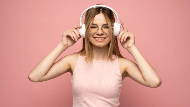 Beautiful attractive young blond woman wearing pink t-shirt and glasses in white headphones listening music and smiling on pink background in studio. Relaxing and enjoying. Lifestyle. - Photo, Image
