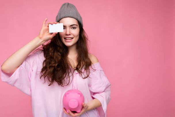 美しい肯定的な陽気なかわいい笑顔若いブルネットの女性の肖像画でスタイリッシュなシャツでピンクの背景に隔離されましたコピースペースと保持ピンクの豚のお金ボックスとクレジットカードのためにモックアップ - 写真・画像