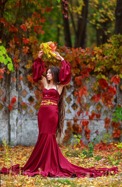 Young lady in a luxurious long burgundy dress with a crown on her head in a colorful autumn park - Fotó, kép