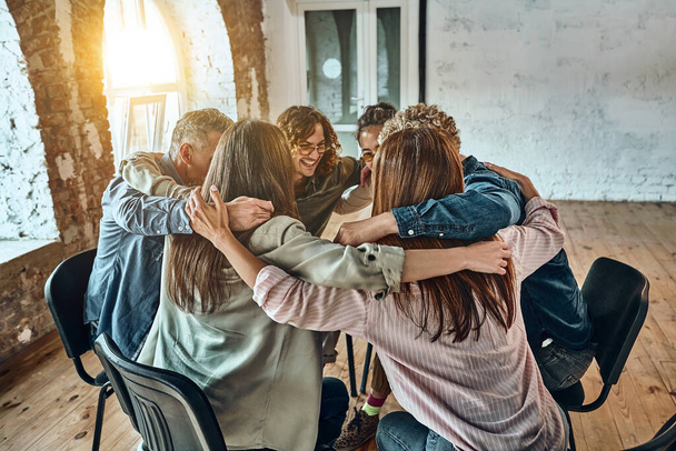 Young people sitting and hugging each other in corporate team building meetings or in group therapy sessions. Concept of mutual support, unity, teamwork, a community of coworkers and business cooperation. Sunbeam background. - Photo, image