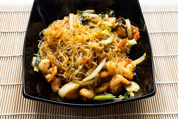 egy adag Mien Xao vietnami wok kevert sült üvegtészta zöldségekkel és fűrészes garnélarák fekete tálban, közvetlen a fa asztalon - Fotó, kép
