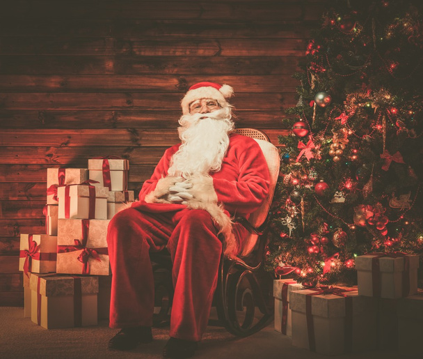 Père Noël assis sur chaise à bascule dans l'intérieur de la maison en bois avec des boîtes-cadeaux autour de lui
  - Photo, image