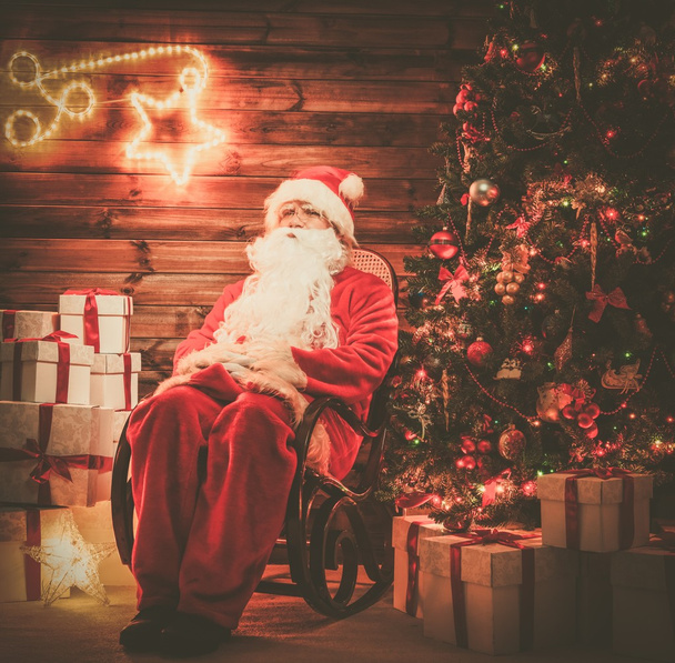 Papai Noel sentado em cadeira de balanço em interior de casa de madeira com decoração iluminada em uma parede
 - Foto, Imagem