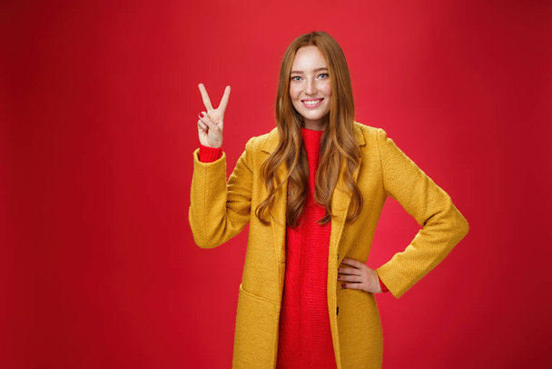 Portrait of happy playful and cute redhead woman with freckles in yellow coat holding hand on waist confident and self-assured showing peace or two sign delighted, beating weather conditions - Photo, Image