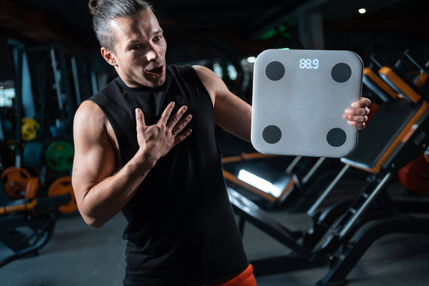 shocked and frustrated man weighing with scale in gym after dieting - Foto, Imagen