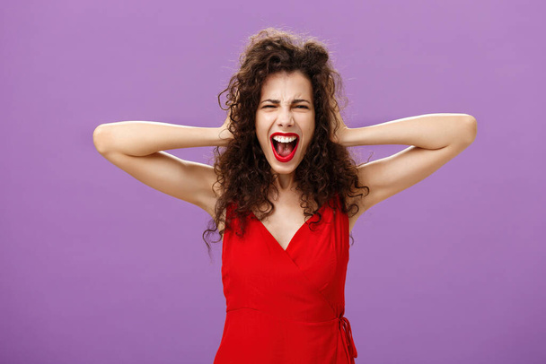 Ex-boyfriend spoilt woman formal event being outraged and pissed yelling in fury holding hands on curly hair grimacing from anger standing over purple background in stylish evening red dress - Foto, imagen