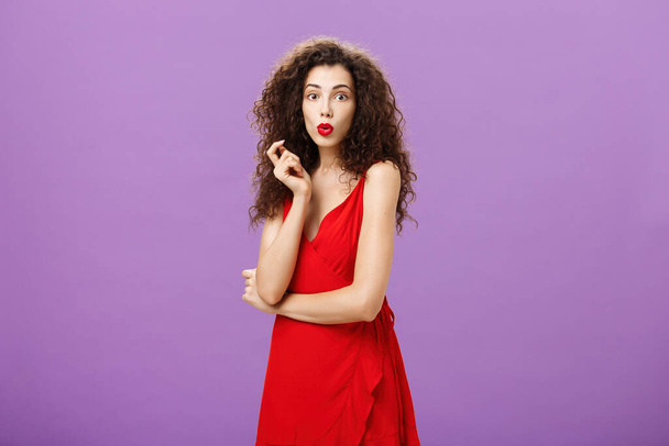 Заінтригована шахраюююча елегантна дама з кучерявою зачіскою в червоній сукні та губками, що розгинаються, дивно торкається волосини, яка вражена чутками, що поширюються навколо вечірки над фіолетовою стіною
 - Фото, зображення