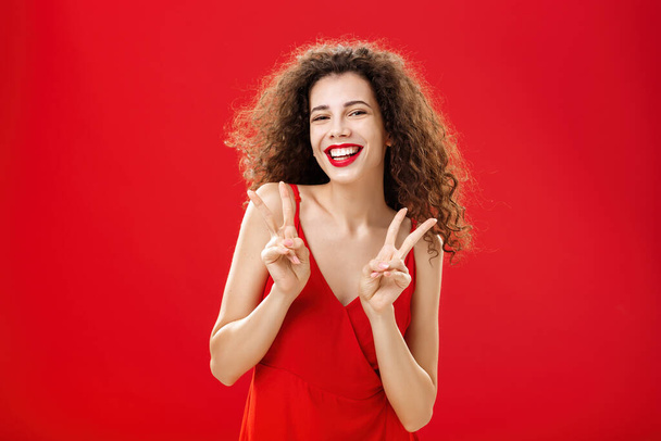 Friendly sociable good-looking happy girl with curly hairstyle wearing elegant red dress and evening make-up showing peace or victory signs joyfully posing over studio background entertained - Fotó, kép