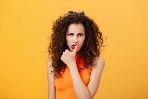Waist-up shot of serious determined and focused displeased woman with curly hairstyle frowning biting thumb looking dangerous and angry standing daring and rebellious over orange background - Photo, Image