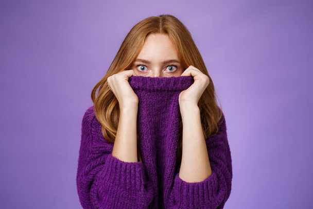 Руда дівчина налякана зима ховається в комірці теплий светр, що вискакує очі шоковані і приголомшені на камеру, ніби наляканий і ненадійний стоячи на фіолетовому фоні
 - Фото, зображення