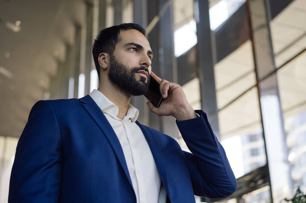 Egy jóképű, töprengő közel-keleti üzletember portréja, aki mobiltelefonon beszél, miközben a modern irodában álló ablakot nézi. - Fotó, kép