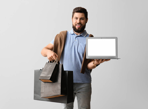 Handsome man with laptop and Black Friday shopping bags on light background - Photo, Image