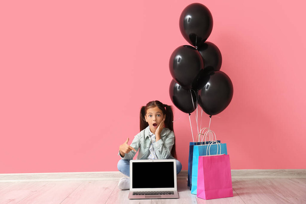 Шокированная маленькая девочка, указывающая на ноутбук возле розовой стены. Чёрная пятница - Фото, изображение